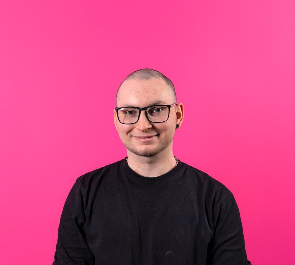 A person in a black t-shirt wearing glasses and smiling with a pink background