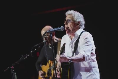 Roger Daltrey, a white man wearing a white short, plays an acoustic guitar while singing into a mic onstage. 