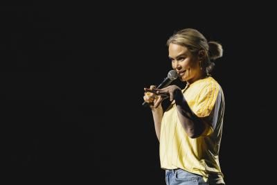 Comedian Joanne McNally onstage at the Royal Albert Hall. She is a blonde woman and is wearing a yellow t-shirt. Her fingers are spread in the air in an expressive gesture. 