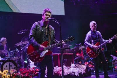Musician Noel Gallagher on stage at the Royal Albert Hall, wearing a leather jacket and playing a guitar. He is surrounded by brightly-coloured flowers 