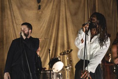 A dramatically-lit shot of Young Fathers on stage at the Royal Albert Hall. On the left is a white man with dark hair and beard wearing a black suit. On the right is a Black man with shoulder-length braids wearing a white shirt. 