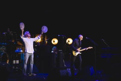 Roger Daltrey, wearing a white shirt and holding two tambourines, stands on stage next to Pete Townshend who is wearing black and playing the guitar