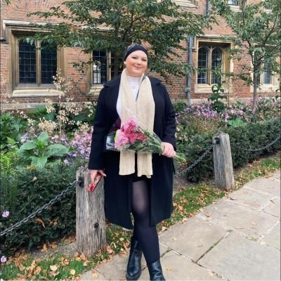 Kirah is smiling whilst holding a bouquet of pink flowers. There is a garden with small trees and purple flowers in the background as well as a red brick building.