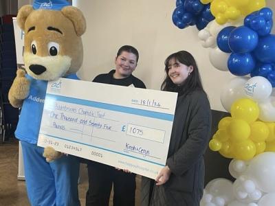 Kirah and her friend Cerys smile holding a big cheque for £1075. Posing beside them, giving a thumbs up, is the Addenbrooke's Hospital mascot, a giant bear wearing blue medical overrals.There is a tall pile of blue, yellow and white balloons in a semi-arch beside them.