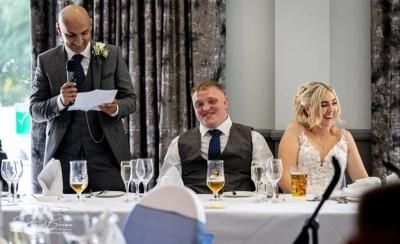 Clayton stands and smiles while reading a best man's speech, the bride and groom are both sat down and laughing beside him.