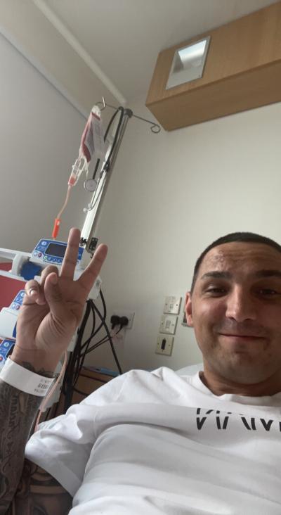 Clayton gives a peace sign to the camera, he's lying back in a hospital bed, you can see treatment machines and IV drip in the background,