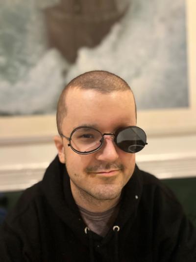 A young man with short hair wearing a black jacket. He is wearing glasses, one lens of which is tinted black.