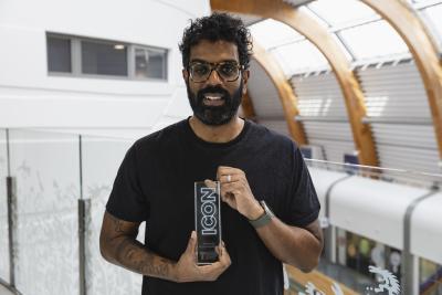 Comedian Romesh Ranganathan is stood with a black top smiling, holding an award which Teenage Cancer Trust gave to him. On the award it says the words Icon